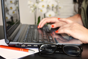 a laptop lying on a desk with hands of a woman typing on the keyboard, a pair of glasses is in front of the picture