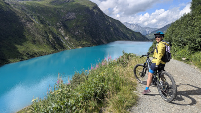a view of a turquoise blue lake with mountains in the background and a mountainbike cyclist posing in front of the picture